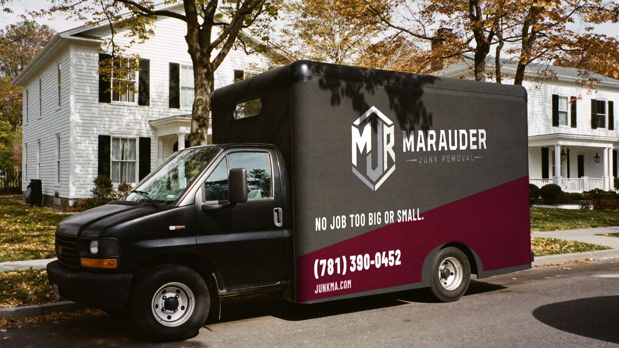 Black Marauder Junk Removal box truck with a maroon band, parked on a leafy street in front of a white colonial house