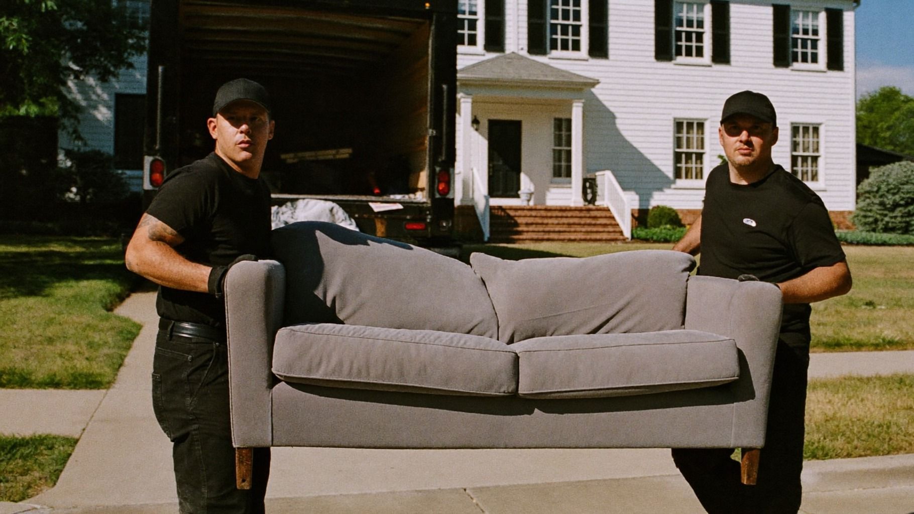 Two crew members in black caps and T-shirts carrying a gray couch down a front walk toward an open box truck