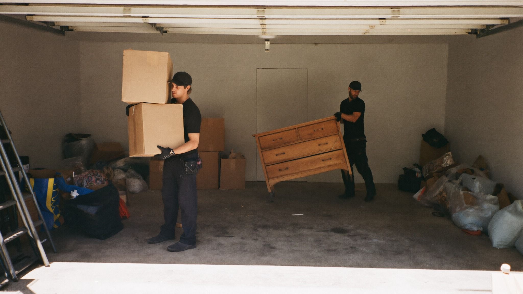 Two crew members clearing a garage, one carrying stacked boxes and the other a wooden dresser